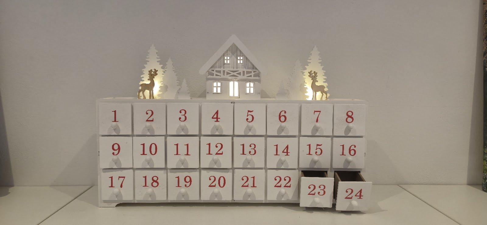 Bílý dřevěný adventní kalendář s šuplíčky a svítícími dekoracemi domečku a sobů na bílém pozadí.